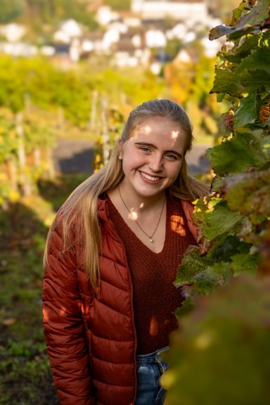 Junge Frau lächelt zwischen Weinreben in einem sonnigen Weinberg.