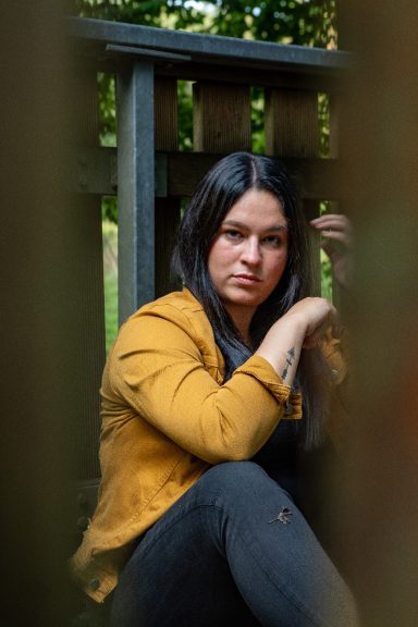 Frau mit langen schwarzen Haaren und gelber Jacke sitzt nachdenklich auf einer Holzplanke.